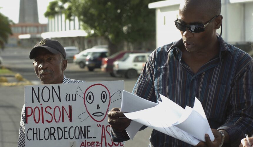 Sur cette photo du 18 novembre 2007, l'avocat Harry Durimel, à droite, se tient à côté d'un militant avec une pancarte indiquant "Non au poison du chlordécone", alors qu'ils protestent lors de l'arrivée de la ministre française de la Santé, Roselyne Bachelot à Raizet Abymes. Durimel fait alors partie des avocats représentant les travailleurs agricoles de Guadeloupe et de Martinique qui luttent depuis longtemps pour obtenir une compensation pour l'utilisation d'un pesticide interdit, le chlordécone. Photo Sipa / AP / Dominique Chomereau-Lamotte